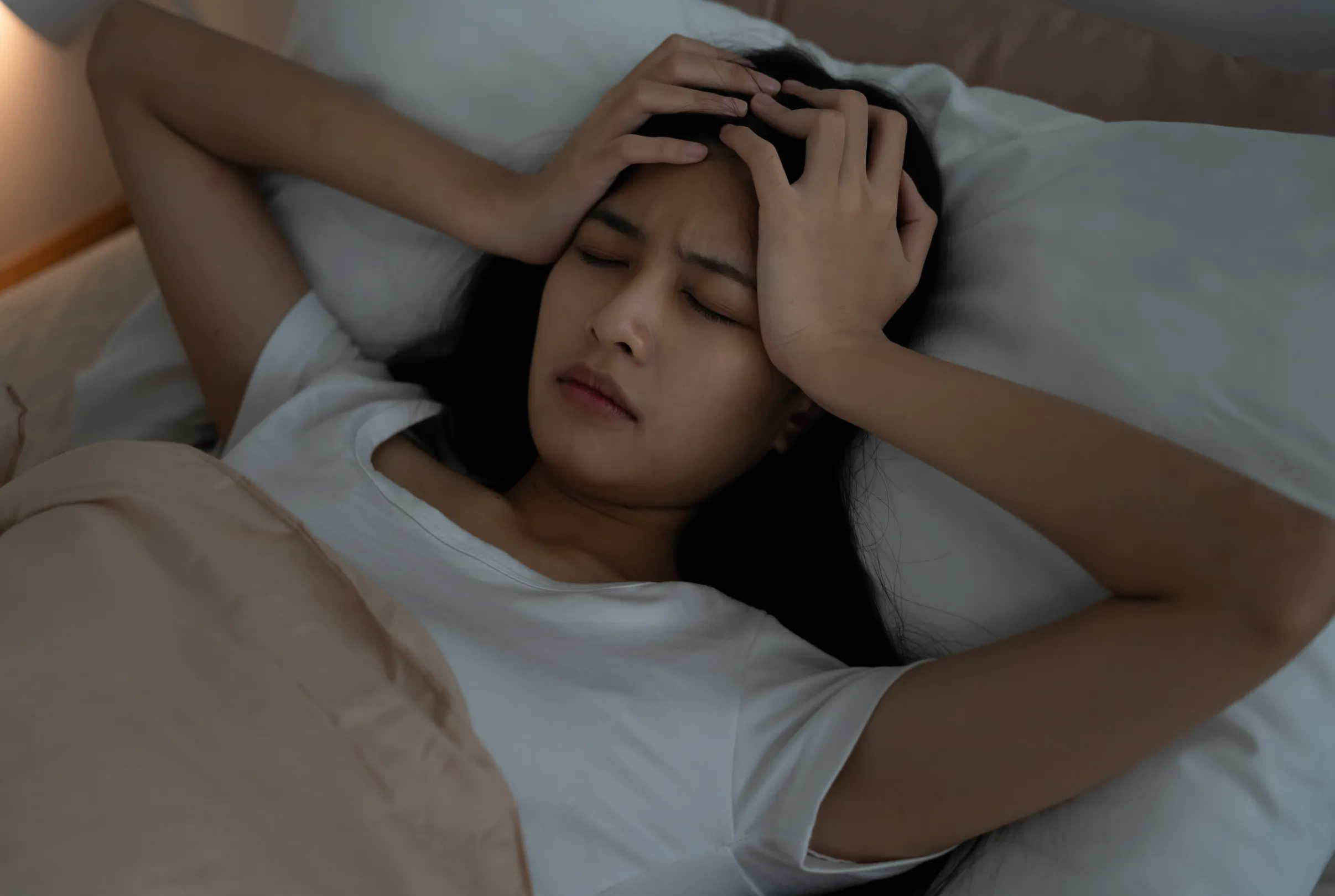 A woman lying in bed at night with her hands pressed against her forehead.