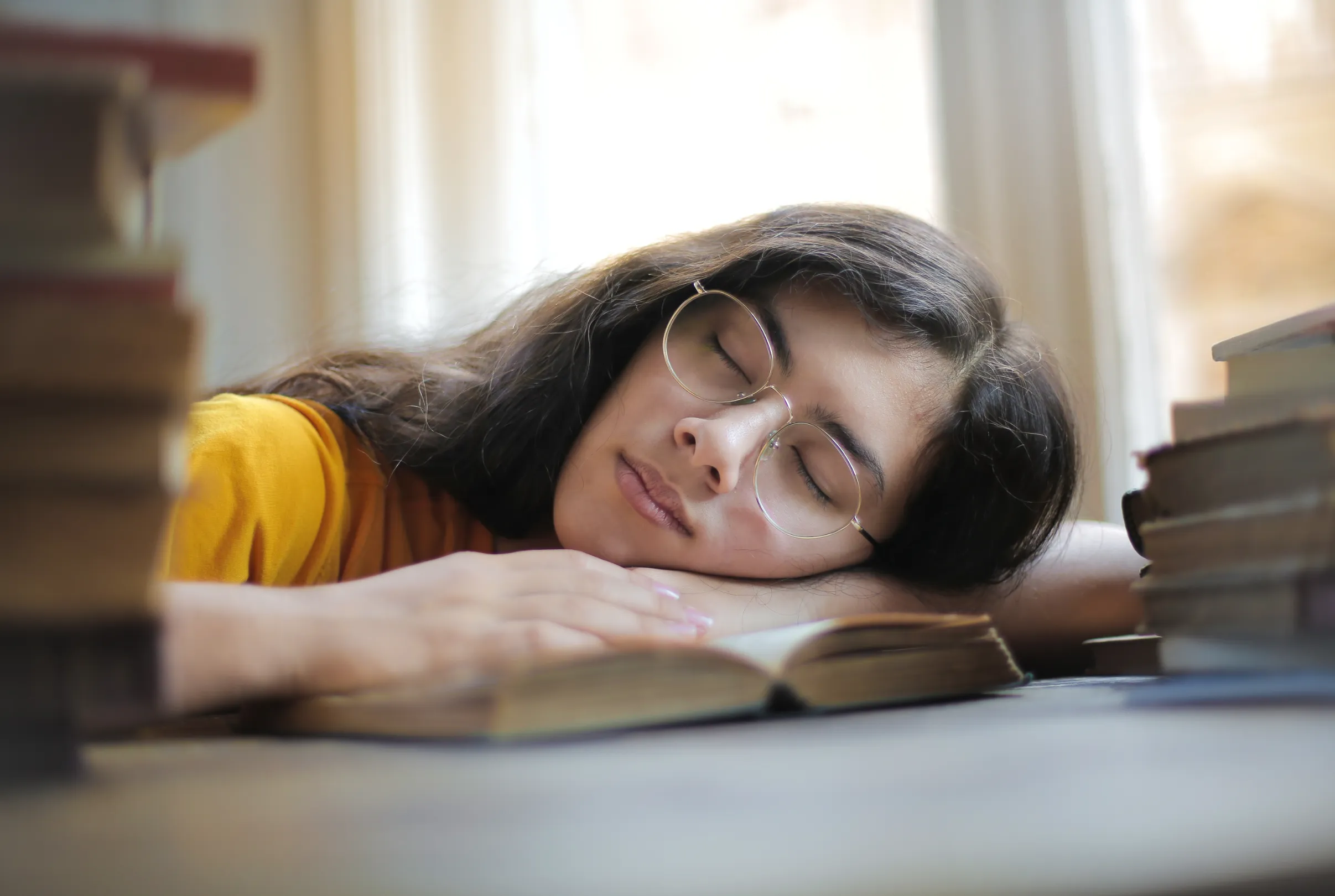 A young woman asleep at a desk with her head resting on an open book, surrounded by stacks of books.