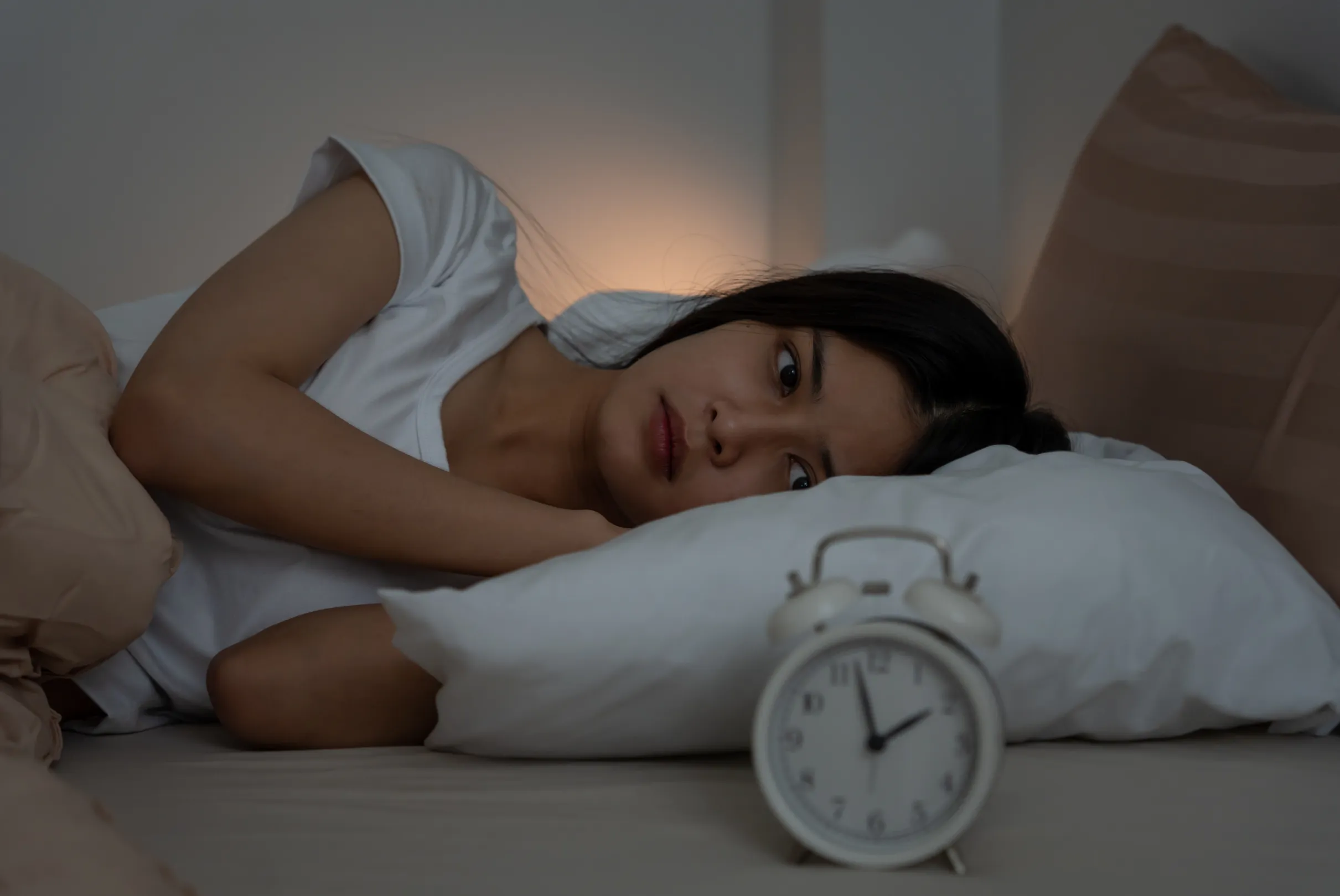 A woman lying awake in a dim bedroom, looking past a white alarm clock on the bedside table.